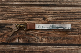 A metal keyring with two keys attached, featuring a silver-toned engraved keychain strap with a blue mountain landscape illustration and a brown leather accent, displayed on a dark wood background.