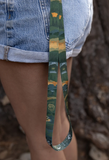 A close-up lifestyle shot of a person wearing light blue denim shorts, showing a patterned green and yellow strap hanging down. The background is a blurry natural outdoor setting with tree bark.
